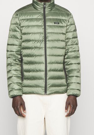 Homme portant une veste matelassée vert clair à fermeture éclair et bordure noire, ainsi qu'un pantalon blanc, debout devant un fond uni.