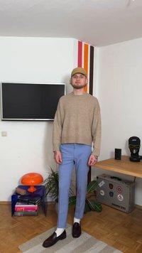 Suéter bege de malha, calças azul claro afuniladas, mocassins escuros com meias brancas. O fundo inclui uma televisão, uma lâmpada laranja e plantas.