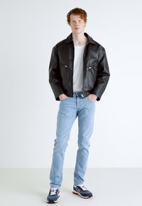 Casaco de pele sintética preto com bolsos com fecho, t-shirt branca, jeans azul claro e ténis azul-marinho com detalhes em vermelho. Modelo está posicionado contra um fundo neutro.