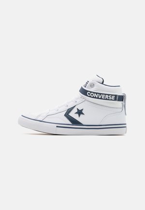 Białe wysokie trampki Converse z granatową gwiazdą w kształcie szewronu, paskiem na kostkę i gumową podeszwą, pokazane z boku na jednolitym tle.