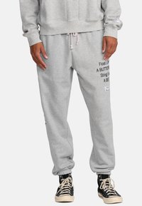 Pantaloni della tuta grigi con polsini elasticizzati, caratterizzati da testo stampato nero sulla gamba sinistra e un piccolo patch del logo vicino alla tasca. Materiale in misto cotone.