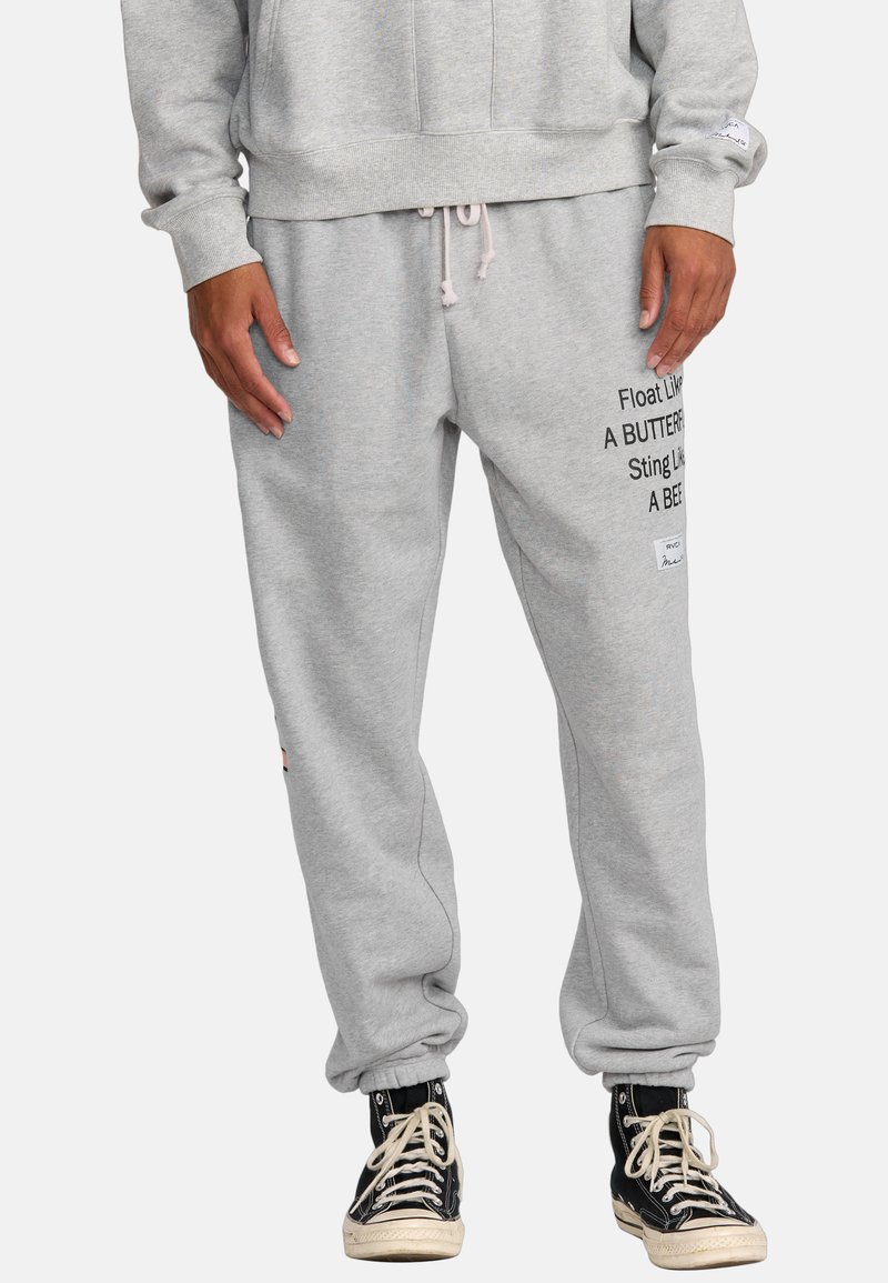 Pantaloni della tuta grigi con polsini elasticizzati, caratterizzati da testo stampato nero sulla gamba sinistra e un piccolo patch del logo vicino alla tasca. Materiale in misto cotone.