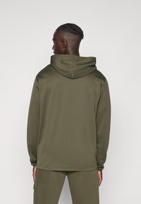 Parka s kapuljačom u maslinasto zelenoj boji, s glatkom teksturom, dugim rukavima i opuštenim krojem. Minimalistički stil bez vidljivih logotipa.