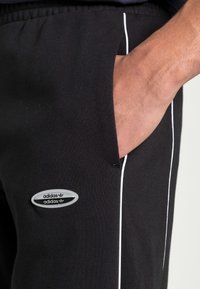 Černé tepláky z bavlny, které mají bílé boční pruhy a oválnou nášivku loga Adidas na levé noze. Elastický pas s kapsami.