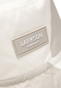 Bežový vedro klobúk vyrobený z hladkej tkaniny s jemným leskom. Obsahuje obdélnikovú, textúrovanú nálepku s nápisom "LARKSON HAMBURG" na bielom pozadí.