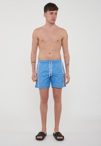 Costume da bagno blu realizzato in tessuto leggero, con chiusura a cordoncino e strisce laterali con testo. Design semplice e taglio alla lunghezza del ginocchio.