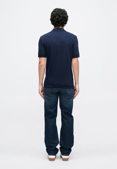 Μπλε σκούρο πόλο με κοντά μανίκια και κλασικό γιακά, συνδυασμένο με σκούρο μπλε τζιν κανονικής γραμμής και ανοιχτόχρωμα αθλητικά παπούτσια, σε θέα από πίσω.