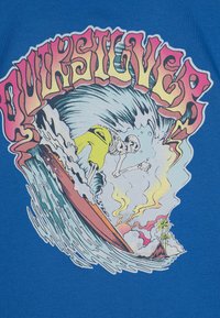 Modrá bavlněná tričko s barevným surfovým motivem, na kterém je kostlivec surfující na vlnách s pozadím západu slunce. Text zní "Quiksilver."