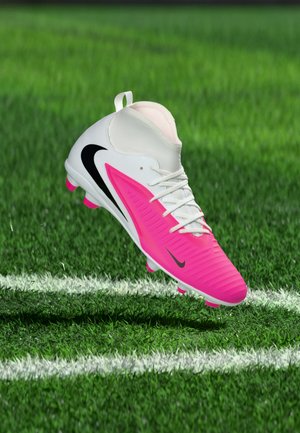 Gheata de fotbal Nike albă și roz plutind deasupra unui teren de iarbă, aproape de linia de delimitare albă, arătând logo-urile negre Swoosh pe lateral și vârf.