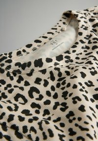 Bluzka w panterkę w kolorze kremowym z czarnymi plamami, z ribbowaną teksturą i okrągłym dekoltem. Widoczna metka z informacjami o pielęgnacji.