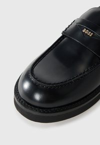 Black leather loafers with a glossy finish, featuring a smooth toe and decorative stitching along the edges, and a branded accent.