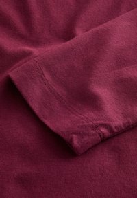 Tricou burgund cu o textură moale, având un tiv al mânecii cu o cusătură curată. Materialul pare a fi de bumbac, cu detalii subtile la suprafață.
