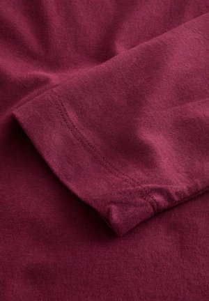 Burgundské tričko vyrobené z měkké tkaniny, se švem rukávu se čistým stehem. Materiál vypadá jako bavlna s jemnými povrchovými detaily.