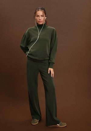 Zelen sweatshirt v oversize kroju z visokim ovratičem in teksturirano tkanino, v kombinaciji z ujemajočimi se širokimi hlačami. Vidne so bele slušalke.