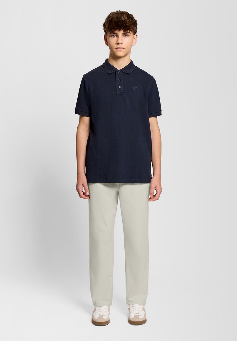 Adolescent debout face à l'avant, portant un polo bleu marine foncé, un pantalon beige clair et des baskets blanches avec des accents beiges.