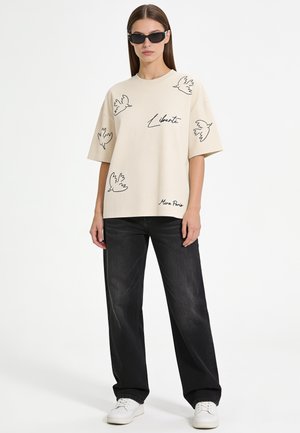Donna che indossa una t-shirt beige oversize con illustrazioni nere di uccelli e testo, jeans neri larghi, sneaker bianche e occhiali da sole neri.