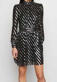 Robe noire avec des rayures métalliques argentées, manches longues et col montant. Taille élastique créant un design plissé pour la jupe.