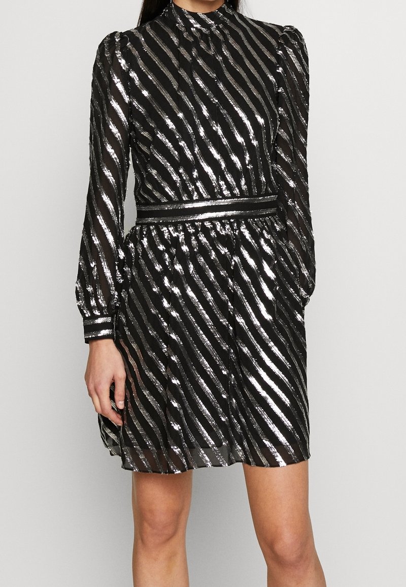 Robe noire avec des rayures métalliques argentées, manches longues et col montant. Taille élastique créant un design plissé pour la jupe.