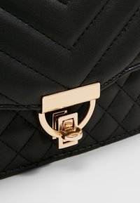 Borsa a tracolla in pelle nera trapuntata con una chiusura geometrica in oro e dettagli. Il materiale presenta un motivo a zigzag con una texture liscia.