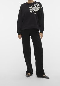 Sudadera negra con bordado floral blanco en el lado izquierdo. Combinada con pantalones anchos negros y zapatos planos puntiagudos negros.