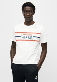 Άσπρο βαμβακερό T-shirt με κεντρικό λογότυπο "CONVERSE ALL STAR" σε μπλε και κόκκινες ρίγες. Μανίκια κοντά και στρογγυλή λαιμόκοψη.