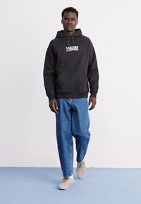 Fekete kapucnis pulóver ezüst logóval, világoskék, laza szabású farmernadrággal és világosbarna sportcipővel párosítva. Egyszerű, hétköznapi megjelenés, minta nélkül.