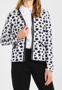 Witte cardigan met marineblauw sterpatroon, contrasterende bies en zakken, gecombineerd met een shirt met kraag en zwarte broek. Zachte stoftextuur.