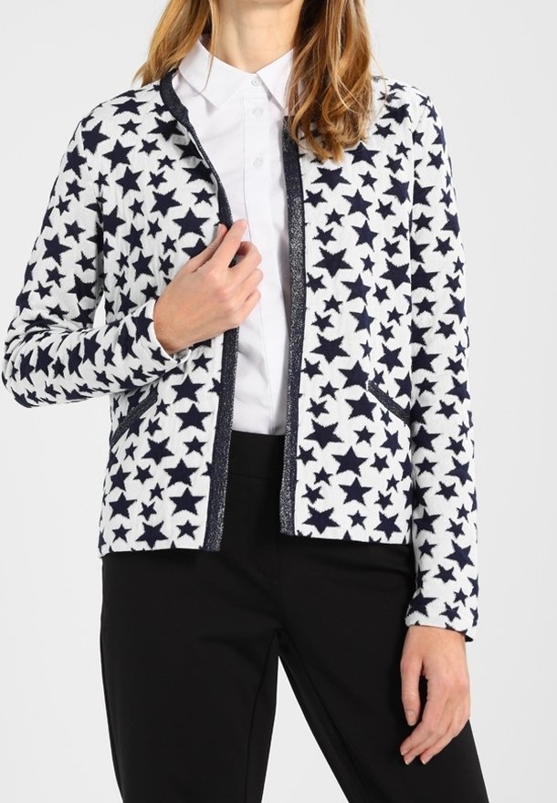 Witte cardigan met marineblauw sterpatroon, contrasterende bies en zakken, gecombineerd met een shirt met kraag en zwarte broek. Zachte stoftextuur.