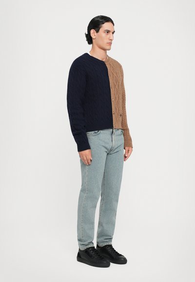 Tvåfärgad kabelstickad kofta med en marinblå vänster sida och en tan höger sida, kombinerad med ljusblå jeans och svarta sneakers.