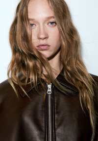 Veste en cuir marron avec une fermeture éclair à l'avant, un petit col et une texture lisse. Le modèle a les cheveux ondulés et une expression neutre.