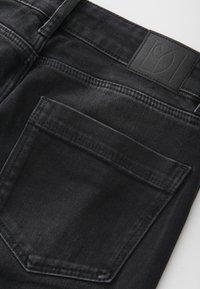 Gros plan sur un jean en denim noir montrant une poche arrière et une étiquette de marque en cuir cousue près de la taille.
