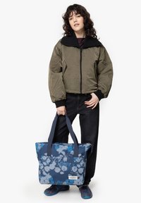 Sac fourre-tout bleu et blanc tie-dye avec deux poignées, en matériau textile, fermeture éclair, poche avant, associé à une veste bomber vert olive.