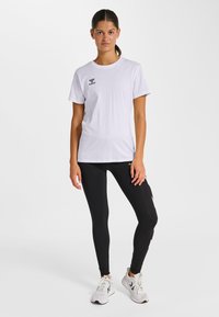 Femme debout devant un fond blanc, portant un t-shirt blanc à manches courtes, des leggings noirs avec des logos sur les côtés, et des baskets blanches.