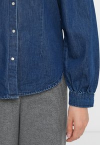 Camisa de ganga em azul profundo com frente abotoada, mangas longas que se ajustam nos punhos, textura suave e bainha arredondada.