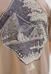 Brun T-shirt med et tekstureret marineblåt broderet bjerglandskab, der indeholder detaljerede linjer og mønstre. Pyntet med et logo nederst.