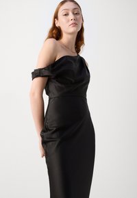 Robe noire décolletée, fabriquée en satin lisse, présentant une silhouette ajustée et un drapé subtil le long de la couture latérale.