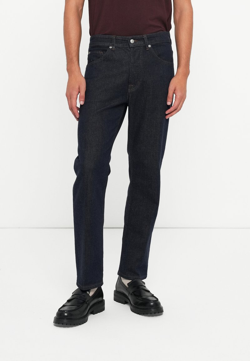 Jeans din denim închis, cu croi drept, cinci buzunare și sistem de închidere cu nasture metalic. Materialul are o textură netedă cu o ușoară decolorare.