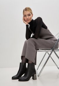 Botas de cuero negras hasta el tobillo con punta cuadrada y tacón bloque texturizado y reflectante. El modelo lleva un ajustado suéter negro de cuello alto y un mono gris con relieve.