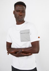 Hvid kortærmet t-shirt med en grå klaplomme, der har et farverigt logo på ærmet og en glat bomuldsstruktur.