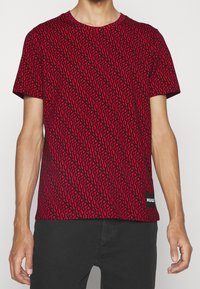 Camiseta roja de manga corta con un patrón gráfico de formas y puntos negros. Presenta una etiqueta negra con "HUGO" en el dobladillo.