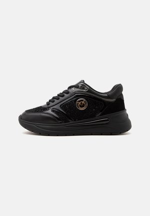 Zapatilla deportiva negra de corte bajo con tejido texturizado y paneles de cuero, cordones negros, suela gruesa y logo circular dorado en el lateral.