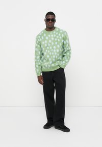 Groene trui met lichtblauwe stippen, geribbelde halslijn en manchetten, gecombineerd met zwarte wijde broek en zwarte sneakers.