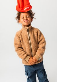 Παιδί που φορά ένα μπεζ μπουφάν από φλις και μπλε παντελόνι ισορροπεί ένα πορτοκαλί πλεκτό καπέλο ανάποδα στο κεφάλι του με κλειστά μάτια και ενώ τα χέρια του είναι ενώμενα.