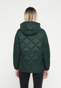 Grønn quiltet jakke med hette, med diamantmønster, elastiske mansjetter og en avslappet passform. Baksidevisning, ingen synlig festemidler.