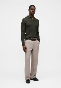 Maglione verde oliva a maglia con scollo a V, abbinato a pantaloni beige chiaro e cintura marrone. Il modello è in piedi con le mani rilassate.