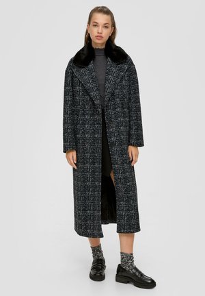 Long navy coat with a textured pattern, black faux fur collar, relaxed fit, and two front buttons. Paired with black shoes and patterned socks.