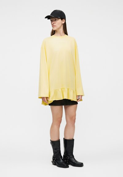 Γυναίκα με φαρδιά κίτρινη φόρεμα με βολάν στο τελείωμα, μαύρη μίνι φούστα, μαύρα μποτάκια και μαύρο καπέλο μπέιζμπολ, στέκεται μπροστά σε λευκό φόντο.