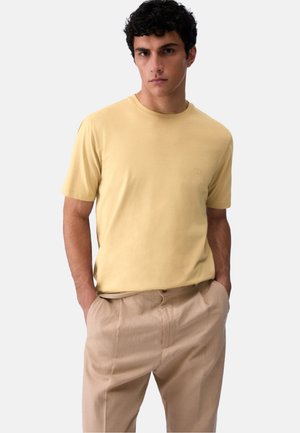 Uomo con capelli scuri e ricci, che indossa una camicia gialla chiara a maniche corte e pantaloni beige, in piedi con le mani nelle tasche davanti a uno sfondo bianco.