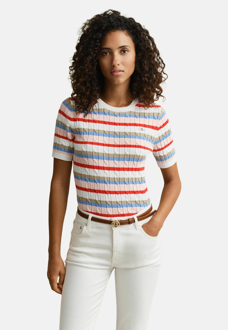 Frau mit lockigem Haar, die ein kurzärmliges, mehrfarbig gestreiftes Oberteil und eine weiße Hose mit braunem Gürtel trägt und mit einer Hand in der Tasche steht.