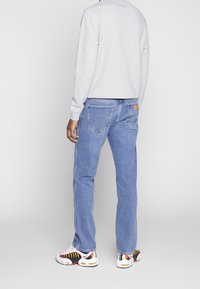 Osoba oblečená v světle šedém svršku, modrých džínech s kapsami vzadu a bílých teniskách s černými, žlutými a červenými akcenty, stojí na šedé podlaze.